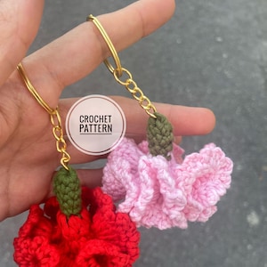 Puede incluir: Dos llaveros de ganchillo con forma de flor de clavel, uno rojo y otro rosa, con tallos verdes y anillas doradas.  "CROCHET PATTERN" está escrito en una etiqueta blanca.