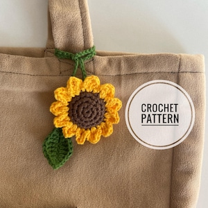 Peut inclure: Un charme en forme de tournesol crocheté avec une feuille verte attachée. Le charme est suspendu à la bandoulière d'un sac fourre-tout brun. Le sac a une poche sur le devant et le texte "CROCHET PATTERN" est écrit dans un cercle sur le sac.