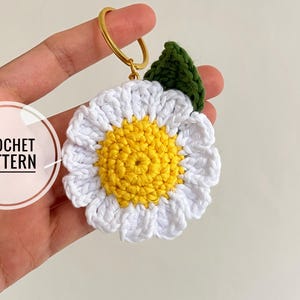 Op de afbeelding: Een handgemaakte gehaakte sleutelhanger in de vorm van een madeliefje met witte bloemblaadjes en een gele kern. De sleutelhanger heeft een groen blad en een goudkleurige ring. De afbeelding bevat de tekst "CROCHET PATTERN" in een witte cirkel.