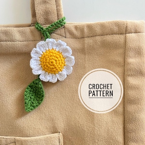 May include: A crochet daisy flower with a green leaf attached to a brown tote bag. The bag has a pocket and the text "CROCHET PATTERN" is written on a white circle.