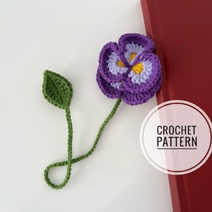 May include: A handmade crochet bookmark shaped like a purple pansy flower with a green stem and leaf. The flower has layers of purple, light purple, and yellow yarn. The bookmark is placed on a red surface.