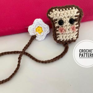May include: A handmade crochet bookmark featuring a slice of toast with a cute face and an egg. The toast has a brown outline and a beige center, with black button eyes and pink cheeks. The egg is white with a yellow center. The text "CROCHET PATTERN" is in a white circle.