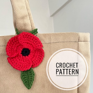 Pode incluir: Uma bolsa tote bege com uma flor de papoula vermelha de crochê presa à alça. A bolsa tem um círculo branco com o texto "CROCHET PATTERN" nele.