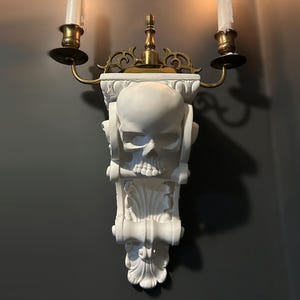 May include: A white skull wall sconce with two candle holders. The candle holders are made of brass and have a traditional design. The skull is detailed and has a realistic look. The sconce is mounted on a wall and has a dark grey background.
