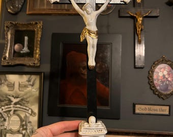 Puede incluir: Un crucifijo de madera negra con una figura de porcelana blanca de Jesús con un taparrabos dorado. La cruz tiene detalles de porcelana blanca en los extremos y una banda con el texto INRI. Se apoya sobre una base de porcelana blanca con calavera.