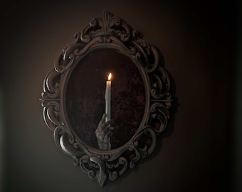 May include: A hand holding a lit candle is reflected in an ornate, dark brown, oval mirror with a snake design.