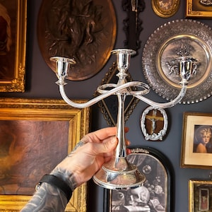 May include: A polished silver-tone metal candelabra featuring a central stem and two curved arms extending outward, each topped with a circular candle cup. The base is circular with a beaded edge detail, and the arms feature a looped design.