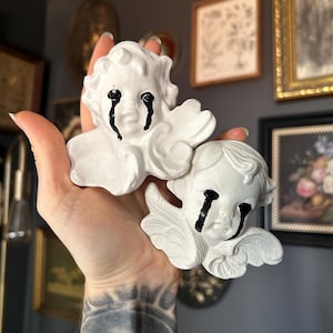 Op de afbeelding: Twee witte cherubijnbeeldjes met zwarte tranenstrepen. De cherubs hebben gedetailleerde vleugels en worden in een hand gehouden. De achtergrond omvat ingelijste kunstwerken en een donkere muur.