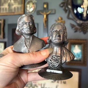 May include: Two silver-toned metal busts depicting historical figures, each mounted on a circular base. The base of the right bust features the engraved text: John Wesley Methodist Bicentennial 1784-1984. Both figures have detailed facial features.