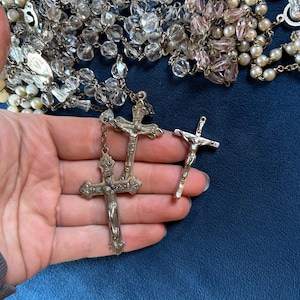 May include: Assortment of rosaries with clear and pink beads, and silver crucifixes. The rosaries feature various bead sizes and shapes. The crucifixes are detailed with the figure of Jesus. The background is a dark blue.