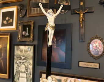 Puede incluir: Un crucifijo de madera negra con una figura de cerámica blanca de Jesús con un taparrabos dorado. En la parte superior hay una banda blanca con las letras INRI. La base es un pedestal de cerámica blanca con una calavera y el texto ECCE HOMO.