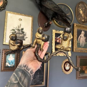 May include: A bronze-toned monkey candle holder with three monkey figures holding bowls. The monkeys are connected by a branch-like structure. The holder is held up by a person's hand. The background includes framed art and a mounted raven.