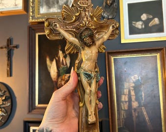 Puede incluir: Un crucifijo de pared que presenta una figura esculpida de Jesucristo en tono beige claro con un taparrabos color verde azulado. La cruz tiene un acabado de pátina dorada y marrón metálica, bordes ornamentados y las letras INRI en la parte superior.