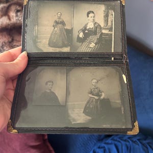 May include: A black leather folding photo album with gold-tone metal corner protectors. The interior features four distinct black and white vintage portrait photographs of individuals in period clothing, displayed behind clear protective plastic sleeves.