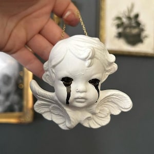 May include: White porcelain cherub ornament with black painted tears, hanging from a gold chain. The cherub has a sad expression and is wearing a white halo.