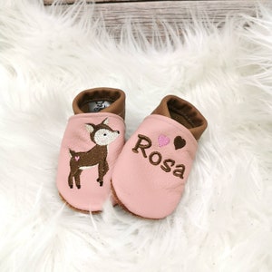 Puede incluir: Un par de zapatos de bebé rosas con ribetes de cuero marrón. Un zapato presenta un ciervo bordado, el otro tiene el nombre "Rosa" bordado con un corazón y un corazón marrón. Los zapatos están sobre una superficie blanca y esponjosa.