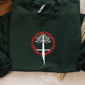 Peut inclure: Un sweat-shirt vert foncé avec un motif d'arbre et d'épée blanc brodé sur le devant. Le motif est entouré d'un cercle rouge avec le texte "Un anneau pour les gouverner tous, Un anneau pour les trouver, Un anneau pour les amener tous, et dans les ténèbres les lier."