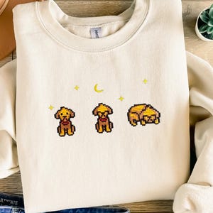Può includere: Una felpa color crema con un design pixelato di tre cani gialli e una luna crescente. I cani sono in pose diverse, uno è seduto, uno è in piedi e uno è sdraiato. Ci sono piccole stelle gialle sparse intorno al design.