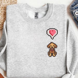 Ash gray unisex crewneck sweatshirt featuring an embroidered Stardew Valley dog character in pixel art style with a small red heart icon above its head. The design reflects the cozy farming game aesthetic with warm brown tones and clean stitching. Casual gamer apparel inspired by Stardew Valley pets, suitable for everyday wear, indie game fans, cozy outfits, and pixel game themed gifting.