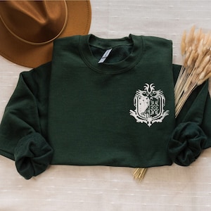 May include: A dark green sweatshirt with a white embroidered design on the left chest. The design features a shield with a wolf and a bird.