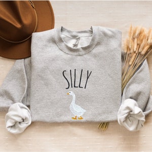 May include: Gray sweatshirt with the word "Silly" printed in black and a white goose illustration.