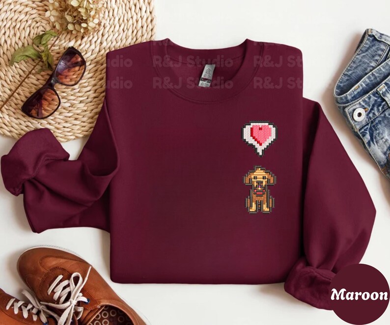 Maroon unisex crewneck sweatshirt showcasing a pixel art embroidered Stardew Valley dog with a red heart icon. The contrast stitching enhances the classic farming game aesthetic, making it a cozy gamer apparel choice for Stardew Valley fans, relaxed streetwear looks, indie game inspired outfits, and thoughtful gaming gifts.