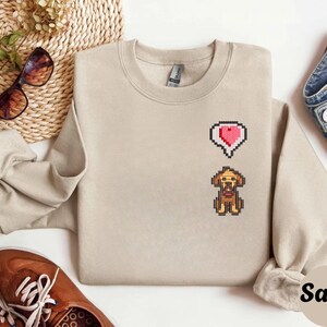 Sand unisex crewneck sweatshirt showcasing a pixel art embroidered Stardew Valley dog with a red heart icon. The contrast stitching enhances the classic farming game aesthetic, making it a cozy gamer apparel choice for Stardew Valley fans, relaxed streetwear looks, indie game inspired outfits, and thoughtful gaming gifts.