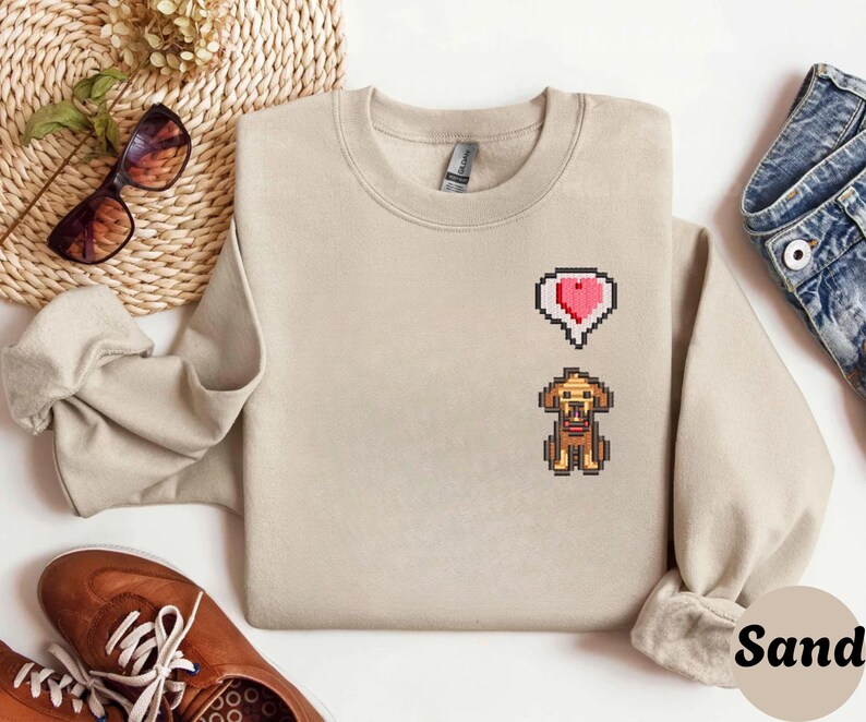 Sand unisex crewneck sweatshirt showcasing a pixel art embroidered Stardew Valley dog with a red heart icon. The contrast stitching enhances the classic farming game aesthetic, making it a cozy gamer apparel choice for Stardew Valley fans, relaxed streetwear looks, indie game inspired outfits, and thoughtful gaming gifts.
