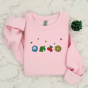 Embroidered Animal Crossing–inspired sweatshirt featuring cute ACNH-style icons stitched on a soft crewneck. Cozy cotton-blend fabric shown in folded and worn views to highlight texture and embroidery detail. Kawaii cottagecore gamer style with pastel color options such as sand, white, blue, and pink. Ideal for cozy gamer outfits, cute embroidered fashion, and cottagecore winter wear. A great gift idea for gamers and fans of soft, comfy embroidered apparel.