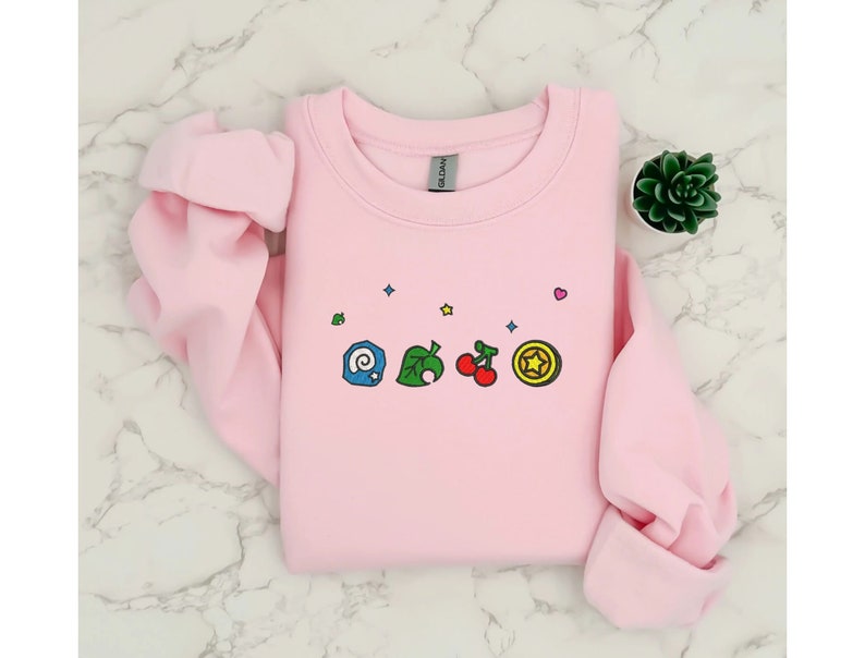 Embroidered Animal Crossing–inspired sweatshirt featuring cute ACNH-style icons stitched on a soft crewneck. Cozy cotton-blend fabric shown in folded and worn views to highlight texture and embroidery detail. Kawaii cottagecore gamer style with pastel color options such as sand, white, blue, and pink. Ideal for cozy gamer outfits, cute embroidered fashion, and cottagecore winter wear. A great gift idea for gamers and fans of soft, comfy embroidered apparel.