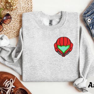 Ash color unisex crewneck sweatshirt featuring a retro sumas bounty hunter helmet embroidery with red armor panels and neon green visor, minimalist gamer streetwear style
Close up sleeve detail showing embroidered retro sumas helmet patch with red stitching and bright green visor on neutral sand crewneck sweatshirt