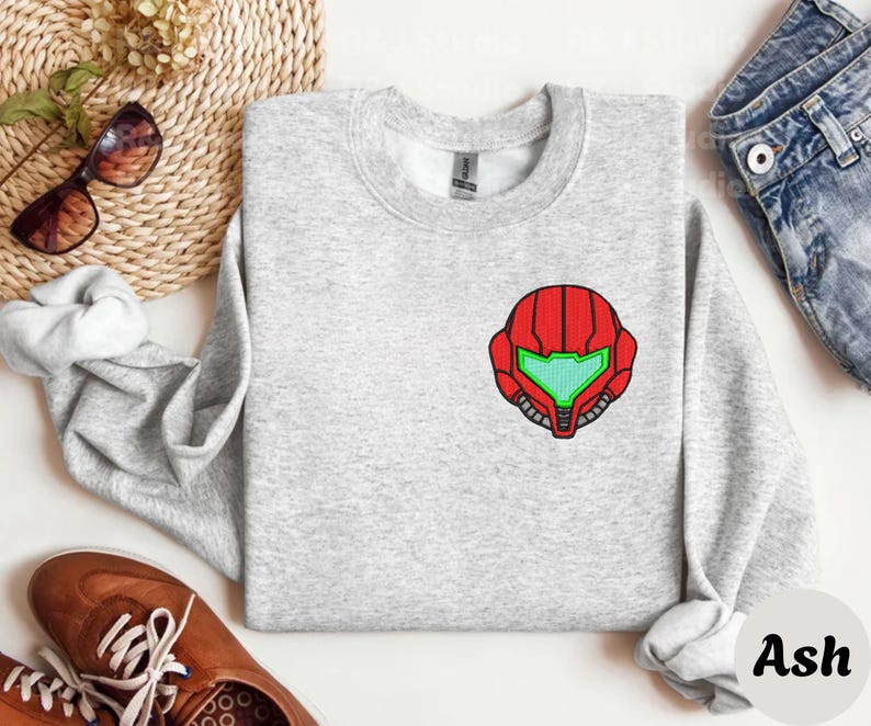 Ash color unisex crewneck sweatshirt featuring a retro sumas bounty hunter helmet embroidery with red armor panels and neon green visor, minimalist gamer streetwear style
Close up sleeve detail showing embroidered retro sumas helmet patch with red stitching and bright green visor on neutral sand crewneck sweatshirt