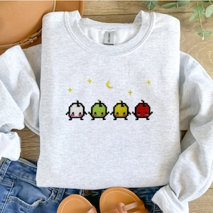 Op de afbeelding: Een grijs sweatshirt met een pixelontwerp van vier cartoonfruitfiguren met een halve maan en sterren op de achtergrond.