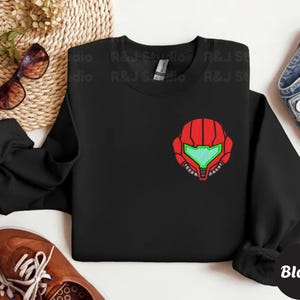 Black color unisex crewneck sweatshirt featuring a retro sumas bounty hunter helmet embroidery with red armor panels and neon green visor, minimalist gamer streetwear style
Close up sleeve detail showing embroidered retro sumas helmet patch with red stitching and bright green visor on neutral sand crewneck sweatshirt
