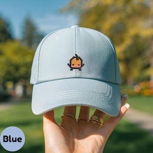 Könnte beinhalten: Hellblaue Baseballkappe mit einem pixeligen orange-braunen Grafikmotiv auf der Vorderseite. Die Kappe wird von einer Hand mit goldenen Ringen gehalten. Das Wort "Blue" befindet sich in einem blauen Kreis unten links.