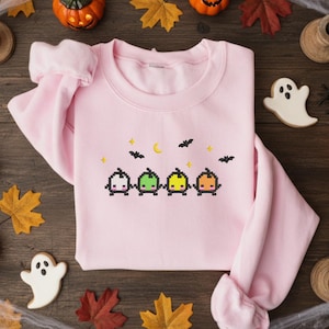 Op de afbeelding: Een lichtroze sweatshirt met een Halloween-thema ontwerp. Het ontwerp toont vier gepixelde pompoenen in verschillende kleuren, met vleermuizen, sterren en een maan erboven. Het sweatshirt is omringd door Halloween-decoraties.