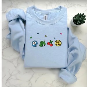 Embroidered Animal Crossing–inspired sweatshirt featuring cute ACNH-style icons stitched on a soft crewneck. Cozy cotton-blend fabric shown in folded and worn views to highlight texture and embroidery detail. Kawaii cottagecore gamer style with pastel color options such as sand, white, blue, and pink. Ideal for cozy gamer outfits, cute embroidered fashion, and cottagecore winter wear. A great gift idea for gamers and fans of soft, comfy embroidered apparel.