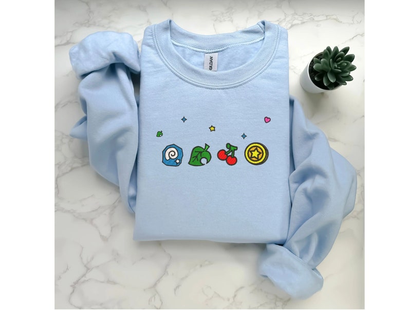 Embroidered Animal Crossing–inspired sweatshirt featuring cute ACNH-style icons stitched on a soft crewneck. Cozy cotton-blend fabric shown in folded and worn views to highlight texture and embroidery detail. Kawaii cottagecore gamer style with pastel color options such as sand, white, blue, and pink. Ideal for cozy gamer outfits, cute embroidered fashion, and cottagecore winter wear. A great gift idea for gamers and fans of soft, comfy embroidered apparel.