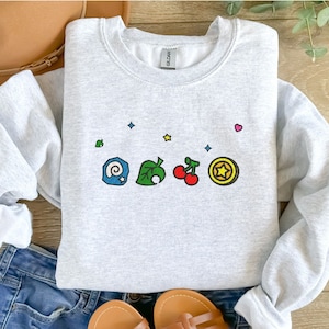 Embroidered Animal Crossing–inspired sweatshirt featuring cute ACNH-style icons stitched on a soft crewneck. Cozy cotton-blend fabric shown in folded and worn views to highlight texture and embroidery detail. Kawaii cottagecore gamer style with pastel color options such as sand, white, blue, and pink. Ideal for cozy gamer outfits, cute embroidered fashion, and cottagecore winter wear. A great gift idea for gamers and fans of soft, comfy embroidered apparel.