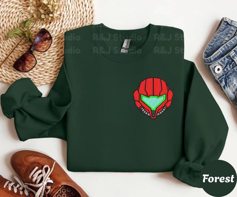 Forest color unisex crewneck sweatshirt featuring a retro sumas bounty hunter helmet embroidery with red armor panels and neon green visor, minimalist gamer streetwear style
Close up sleeve detail showing embroidered retro sumas helmet patch with red stitching and bright green visor on neutral sand crewneck sweatshirt