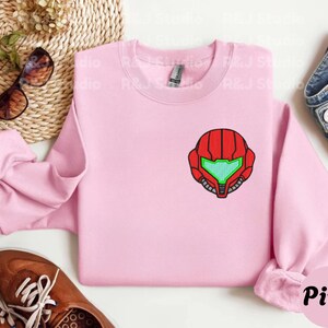 Pink color unisex crewneck sweatshirt featuring a retro sumas bounty hunter helmet embroidery with red armor panels and neon green visor, minimalist gamer streetwear style
Close up sleeve detail showing embroidered retro sumas helmet patch with red stitching and bright green visor on neutral sand crewneck sweatshirt