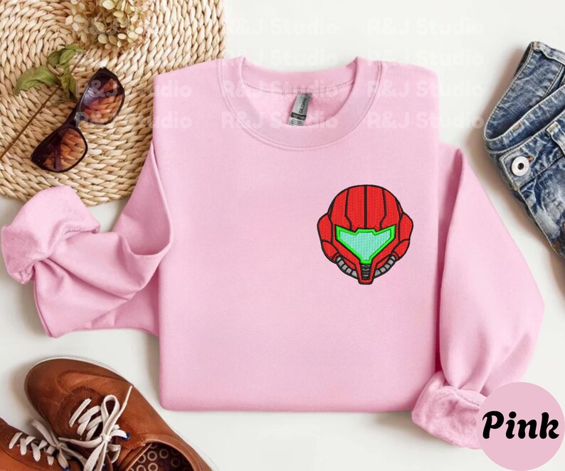 Pink color unisex crewneck sweatshirt featuring a retro sumas bounty hunter helmet embroidery with red armor panels and neon green visor, minimalist gamer streetwear style
Close up sleeve detail showing embroidered retro sumas helmet patch with red stitching and bright green visor on neutral sand crewneck sweatshirt