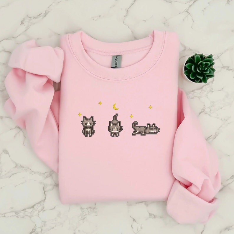 Pu&ograve; includere: Una felpa rosa con un design pixelato di tre gatti e una luna crescente. I gatti sono grigi e la luna e le stelle sono gialle.