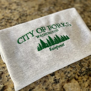 May include: A folded, light gray sweatshirt with green embroidered text and graphics. The text reads "CITY OF FORKS WASHINGTON Rainforest" with a forest graphic below the city name.