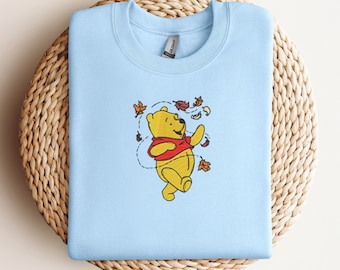 May include: A light blue sweatshirt featuring an embroidered image of a yellow bear in a red shirt, surrounded by falling autumn leaves. The bear is smiling and appears to be dancing. The sweatshirt is displayed on a woven, circular surface.