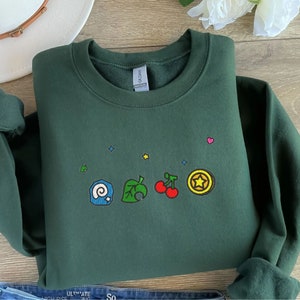 Op de afbeelding: Een donkergroen sweatshirt met een wit, geel, rood, blauw en roze geborduurd ontwerp van verschillende Animal Crossing-personages. Het ontwerp bevat een blad, een bel, een kers en een spiraalvormige schelp.