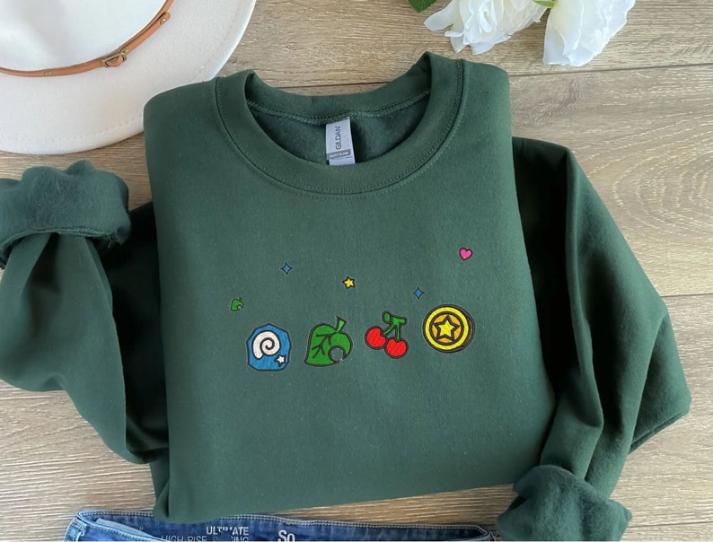Embroidered Animal Crossing–inspired sweatshirt featuring cute ACNH-style icons stitched on a soft crewneck. Cozy cotton-blend fabric shown in folded and worn views to highlight texture and embroidery detail. Kawaii cottagecore gamer style with pastel color options such as sand, white, blue, and pink. Ideal for cozy gamer outfits, cute embroidered fashion, and cottagecore winter wear. A great gift idea for gamers and fans of soft, comfy embroidered apparel.
