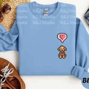 Light blue unisex crewneck sweatshirt featuring a small embroidered Stardew Valley pixel dog with a red heart icon. The design reflects the calming, cozy farming game atmosphere of Stardew Valley, making it suitable for everyday casual wear, indie game lovers, pixel art fans, and comfortable gamer themed clothing.