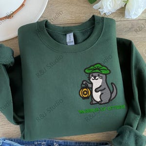 May include: A dark green sweatshirt featuring an embroidered cartoon otter wearing a green hat and holding a lantern. The text "WARRIOR OF OTTERS" is embroidered below the otter. The sweatshirt is a Gildan brand.