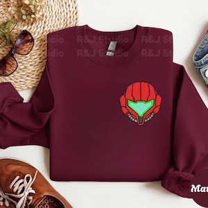 Maroon color unisex crewneck sweatshirt featuring a retro sumas bounty hunter helmet embroidery with red armor panels and neon green visor, minimalist gamer streetwear style
Close up sleeve detail showing embroidered retro sumas helmet patch with red stitching and bright green visor on neutral sand crewneck sweatshirt